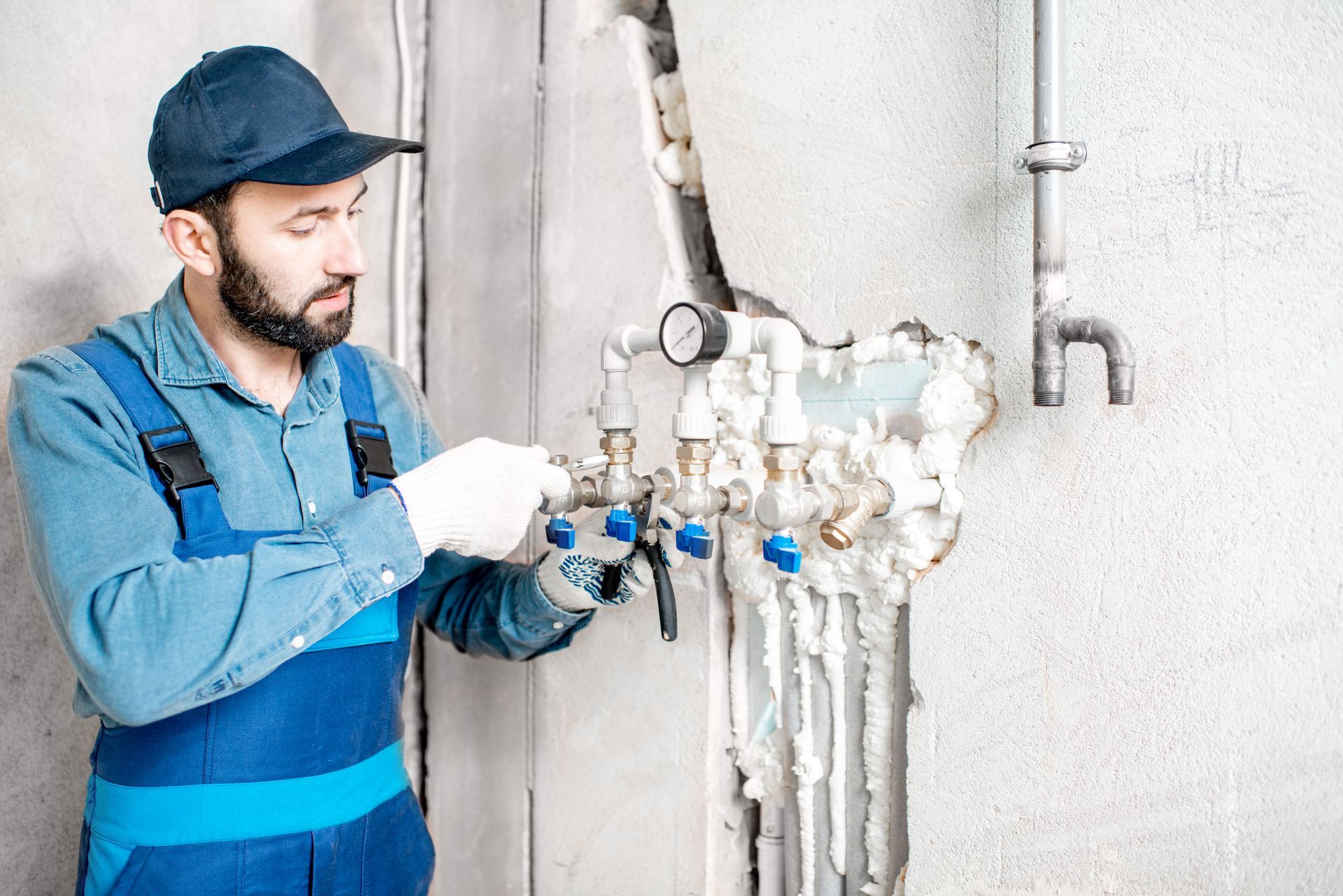 Plumber in blue overalls and cap, working on pipes in a wall.