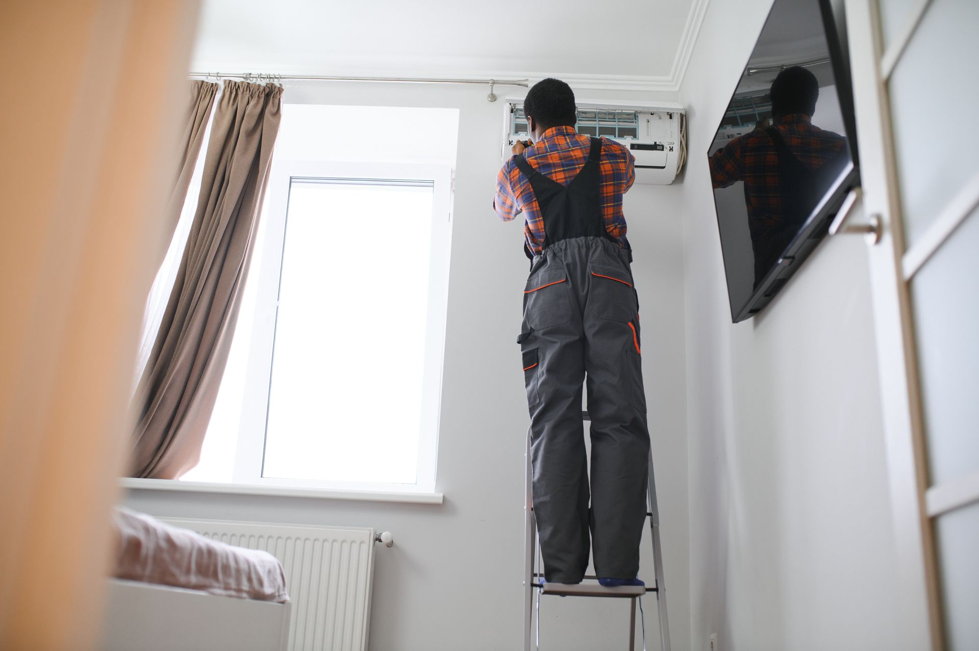 A person in overalls on a ladder fixing an air conditioner near a window and TV.