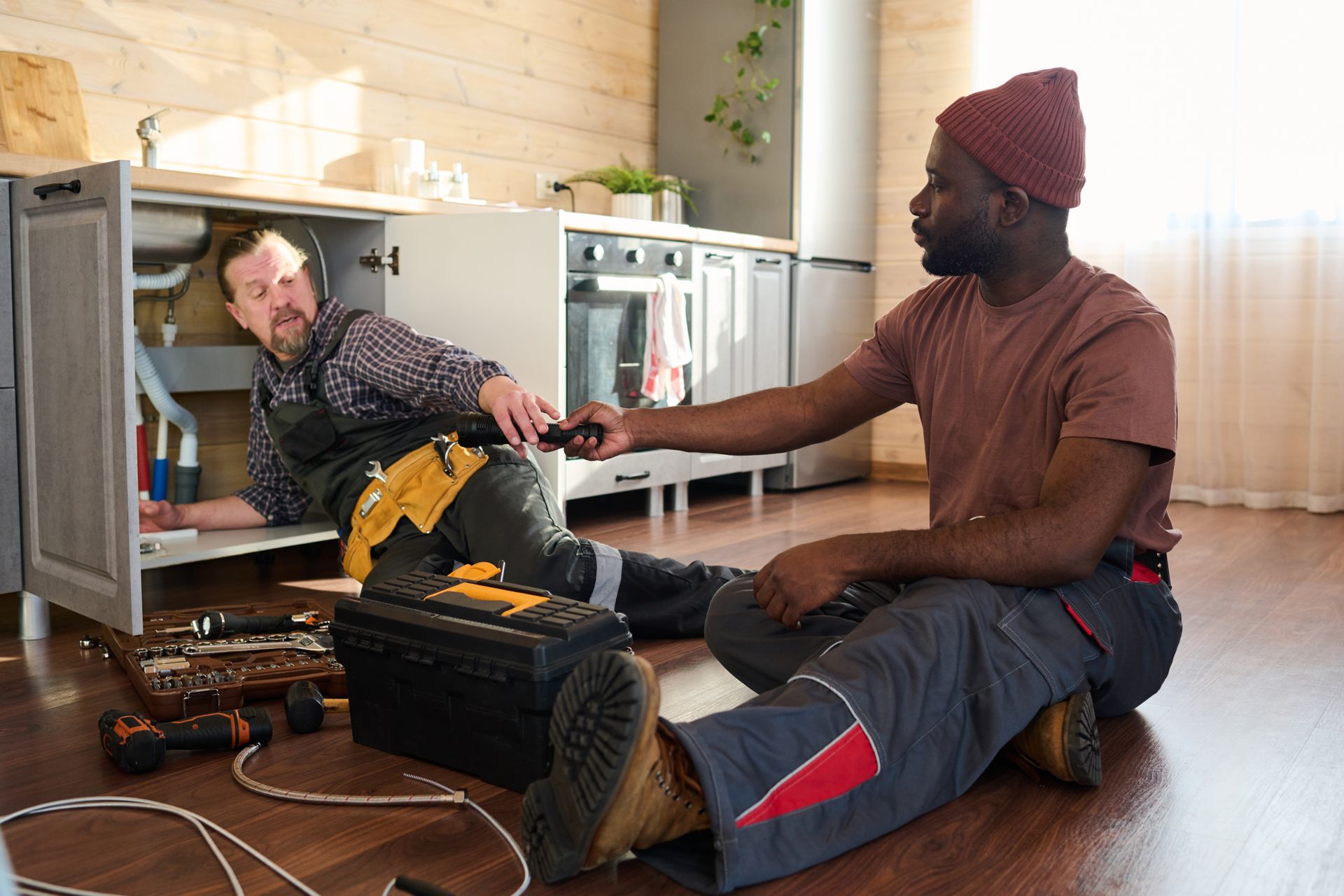 Two men working on plumbing under a kitchen sink; one inside, one handing a tool.