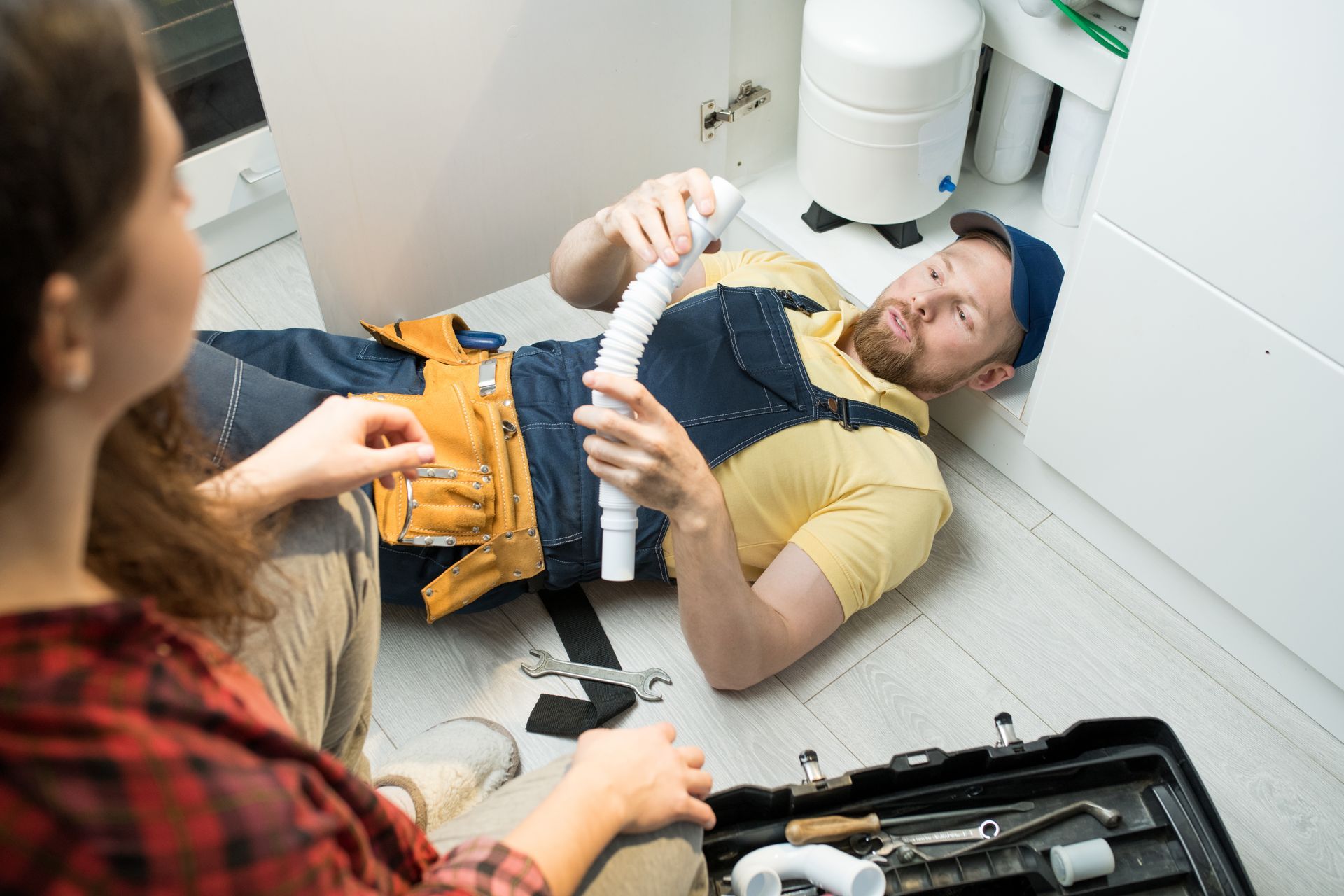 Plumber lying under sink, examining pipe, tool case in foreground, woman looking on.