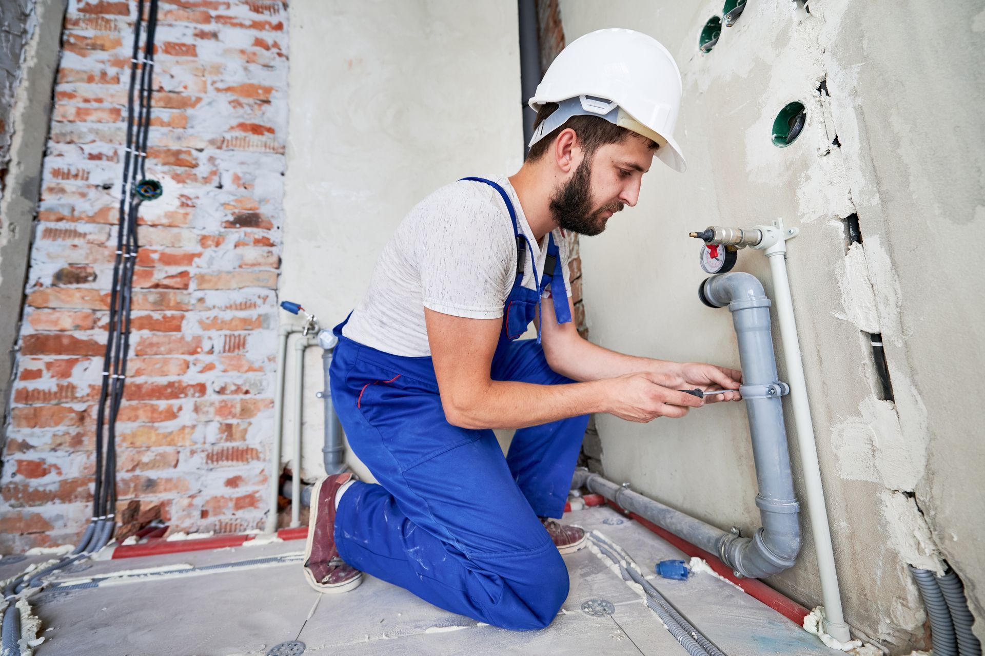Plumber in blue overalls and white hard hat, kneeling and working on pipes in a construction site.