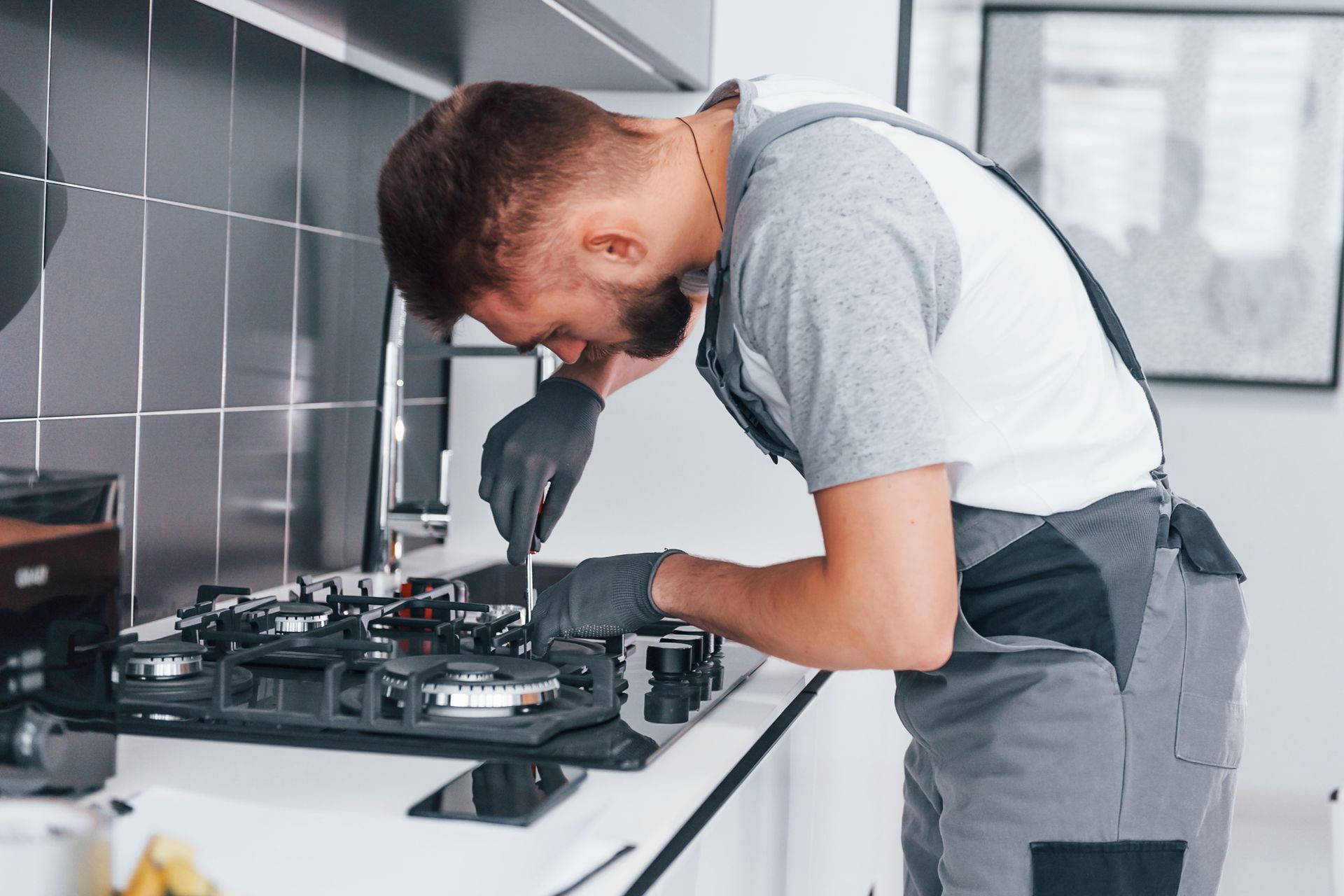Man in work clothes repairing a gas stove in a modern kitchen.