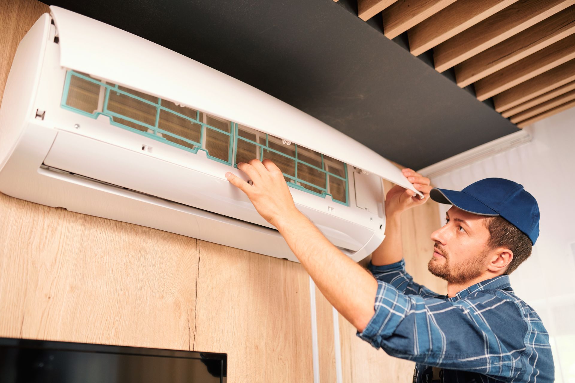 Man in blue cap and plaid shirt cleaning an air conditioner filter indoors.
