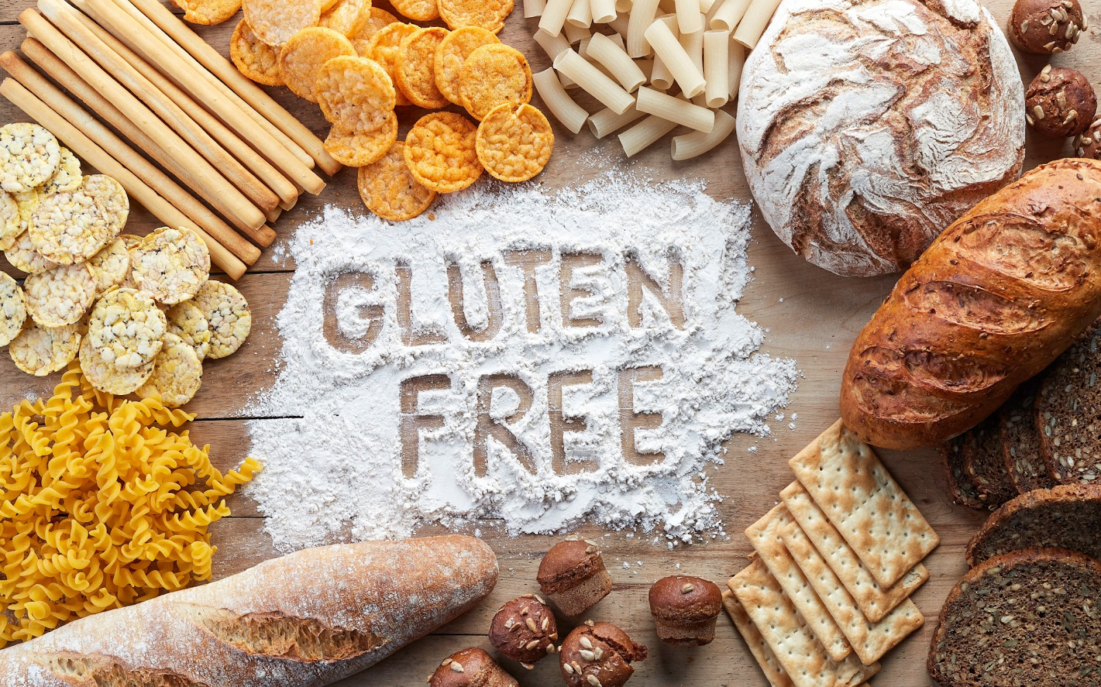 The word gluten free is written in flour on a wooden table surrounded by different types of food.