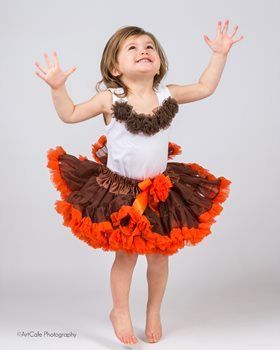Child jumping, wearing a white top and brown and orange tutu, arms raised, smiling.