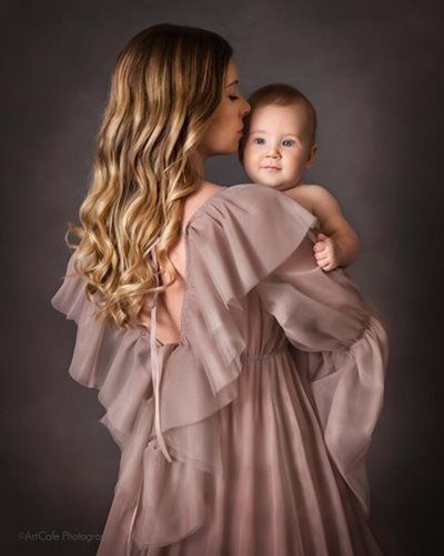 Woman in a flowing mauve gown kisses a baby's head. Dark gray backdrop.