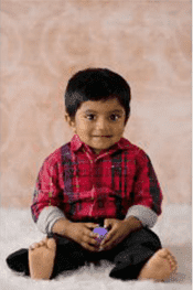 Child sitting, smiling, holding a small purple toy, wearing a red plaid shirt, black pants.