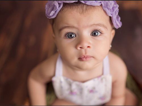 Baby with wide eyes and purple floral headband, wearing a white outfit, looking up.