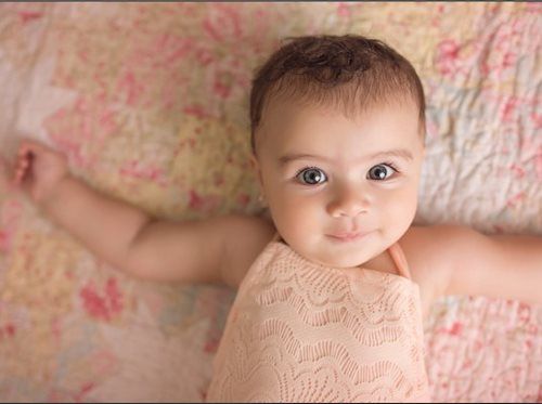 Baby lying on floral patterned blanket, arms outstretched, wearing pink dress, smiling.