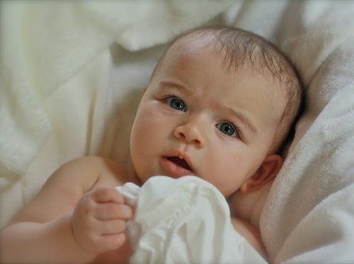 Baby with wide eyes, looking at the viewer. Holding a white cloth, resting in a white blanket.
