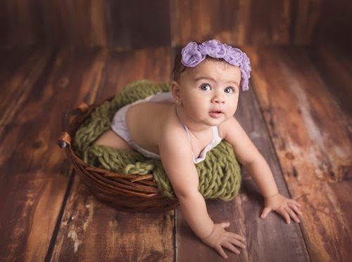 Baby in a basket with green blanket and purple floral headband on a wooden background.
