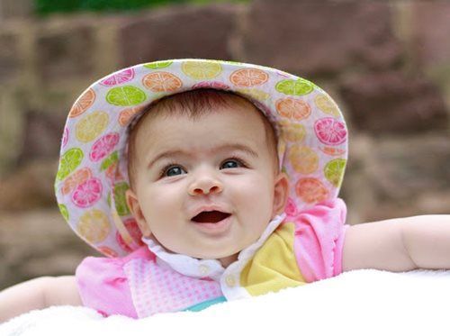 Baby wearing a colorful patterned hat, smiling with open mouth, outdoors.