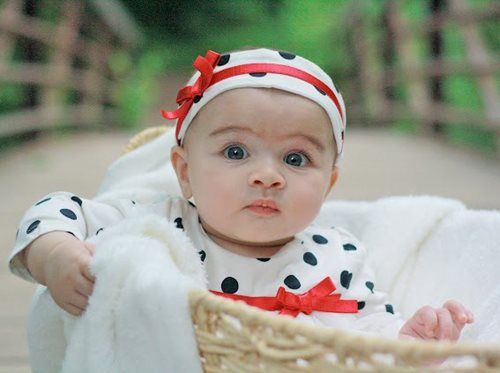 Baby wearing polka dots, seated in a wicker basket, on a bridge, looking at the camera.