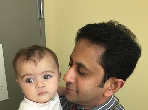 Man holding baby; both looking to the left. Baby has a neutral expression. Bright room, yellow wall.