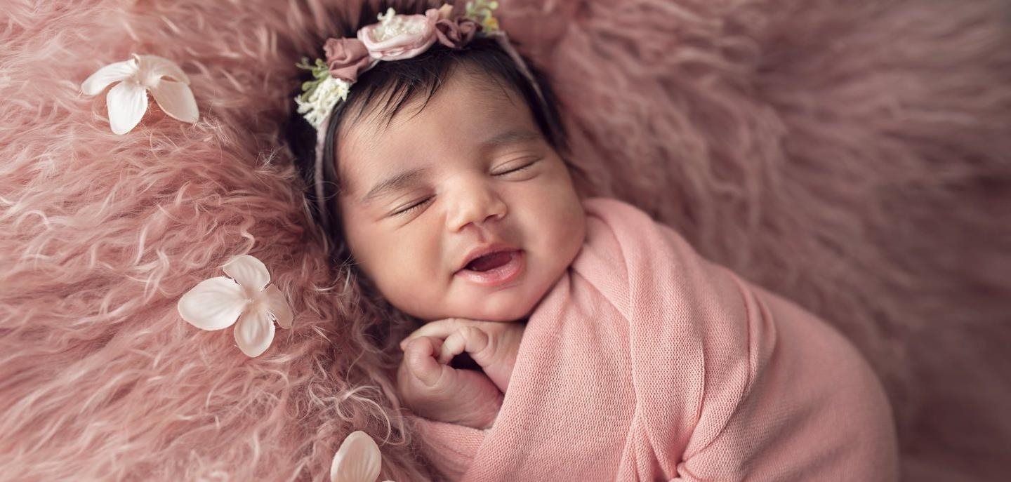 Newborn wrapped in pink with a floral headband, smiling peacefully on a pink fluffy surface, and flowers.