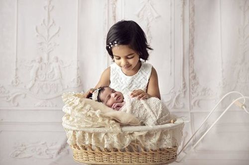 Older child smiles at newborn in a wicker bassinet with cream-colored lace, against a white, ornate wall.