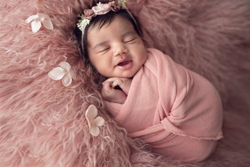 Newborn baby swaddled in pink, smiling, wearing floral headband, on pink fluffy blanket.