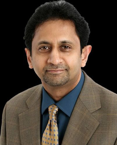 Man in suit, smiling, with a blue shirt and patterned tie against a black background.