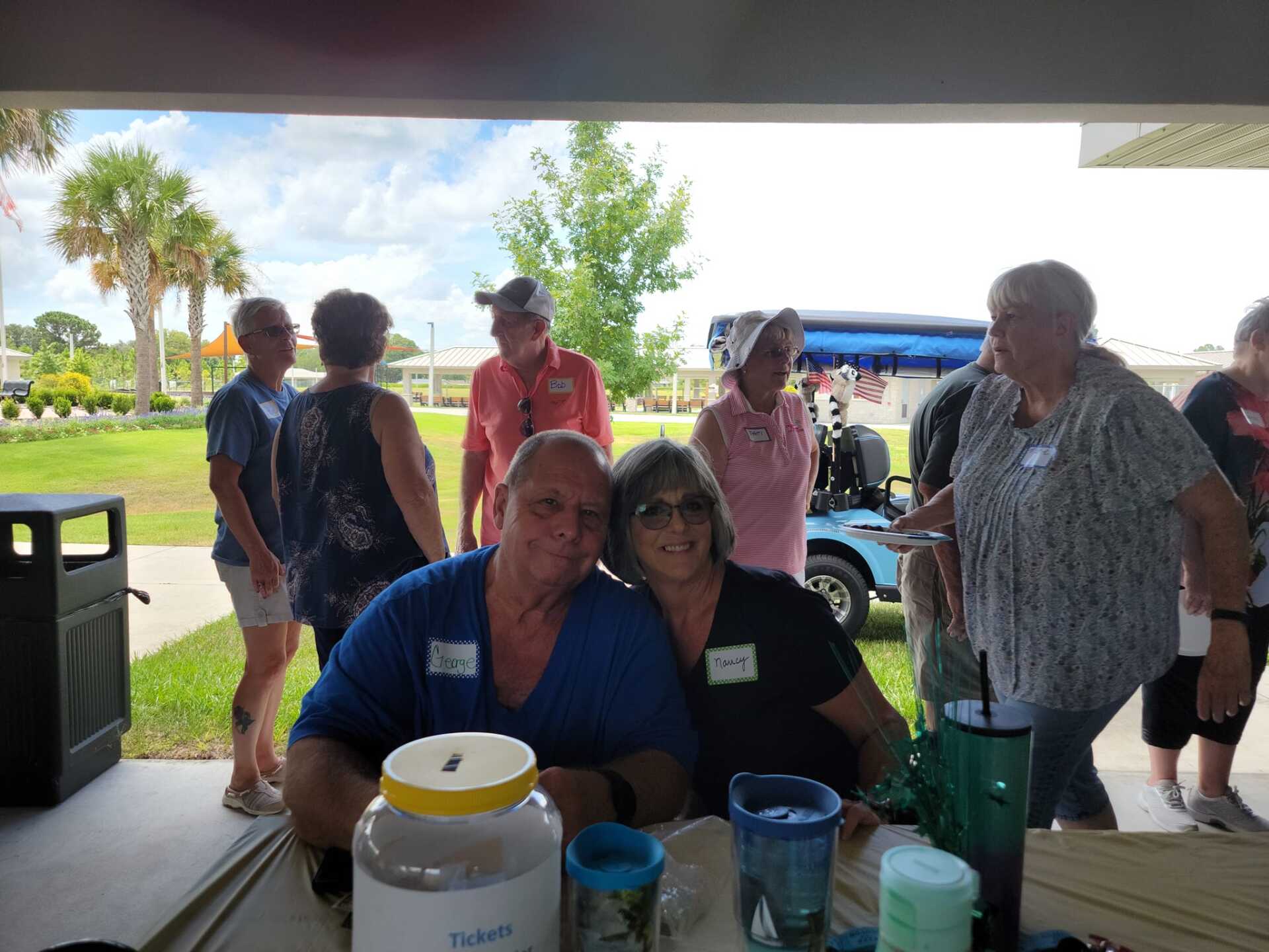 People gathered at an outdoor event, two in foreground smiling, others in background. Sunny day.