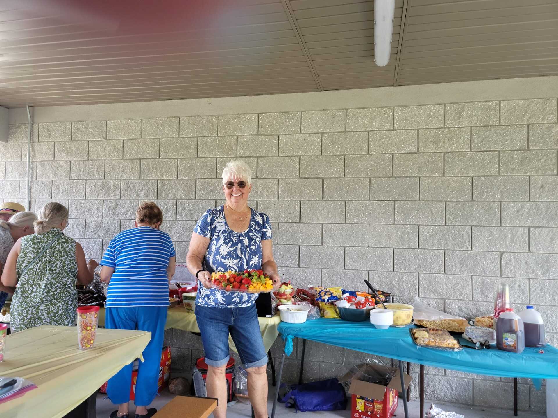 Woman holding a skewer of food smiles at camera at an outdoor party, standing in front of a buffet table.