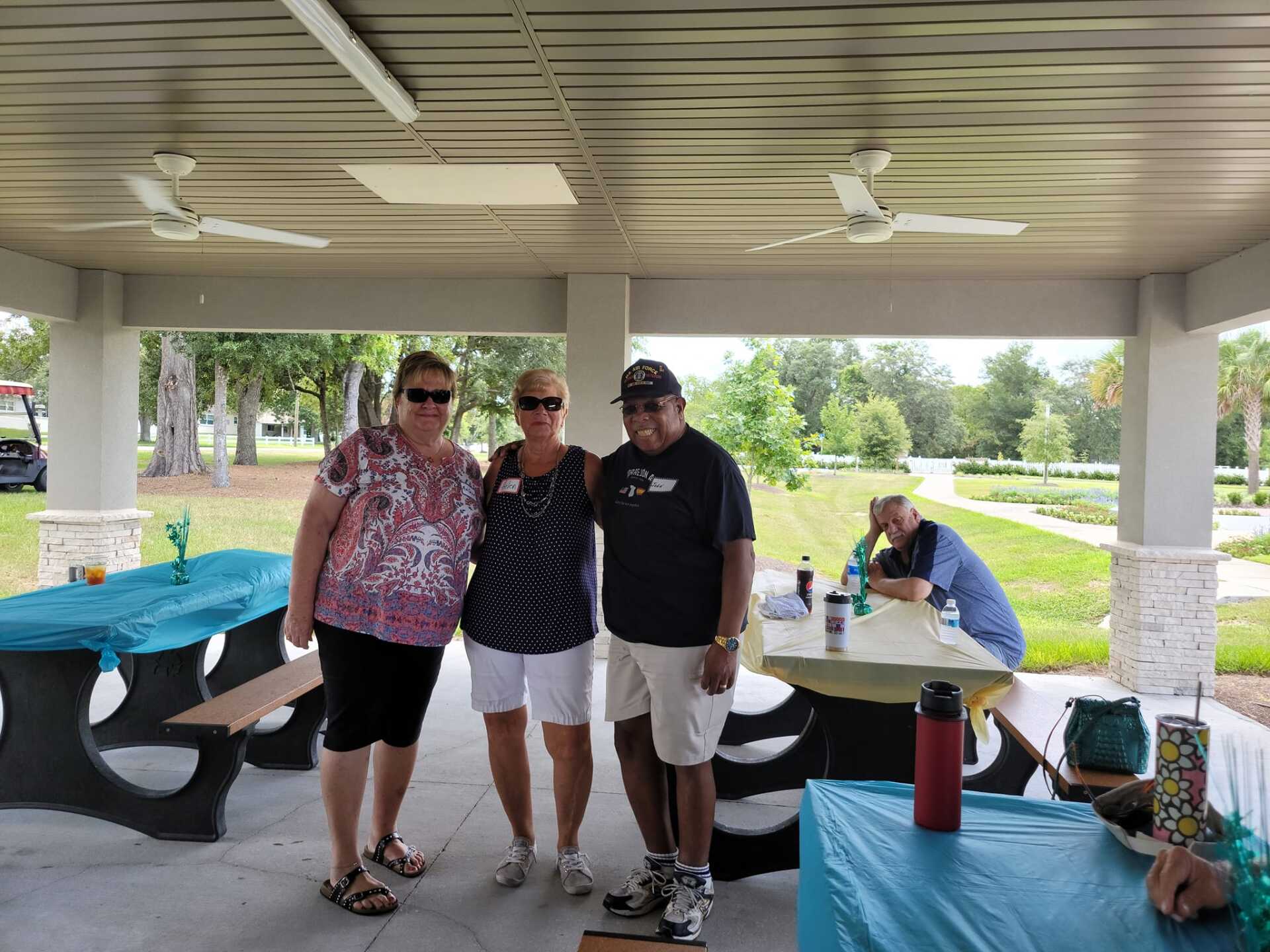 Three people smiling under a covered picnic area. One has arm around another. Turquoise tables. Park in the background.