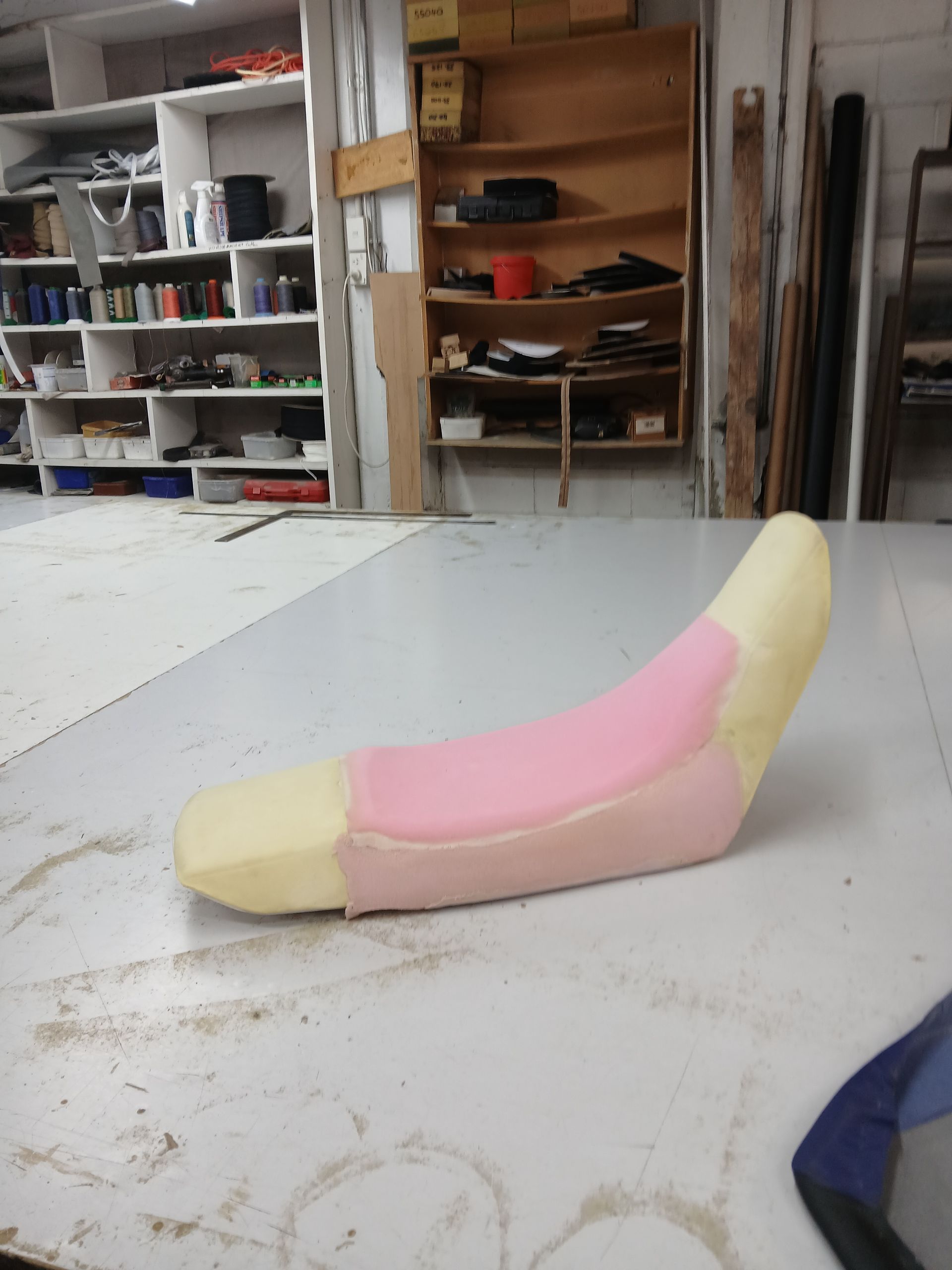 Foam motorcycle seat in workshop, light pink and yellow, on a white surface. Shelves in the background.