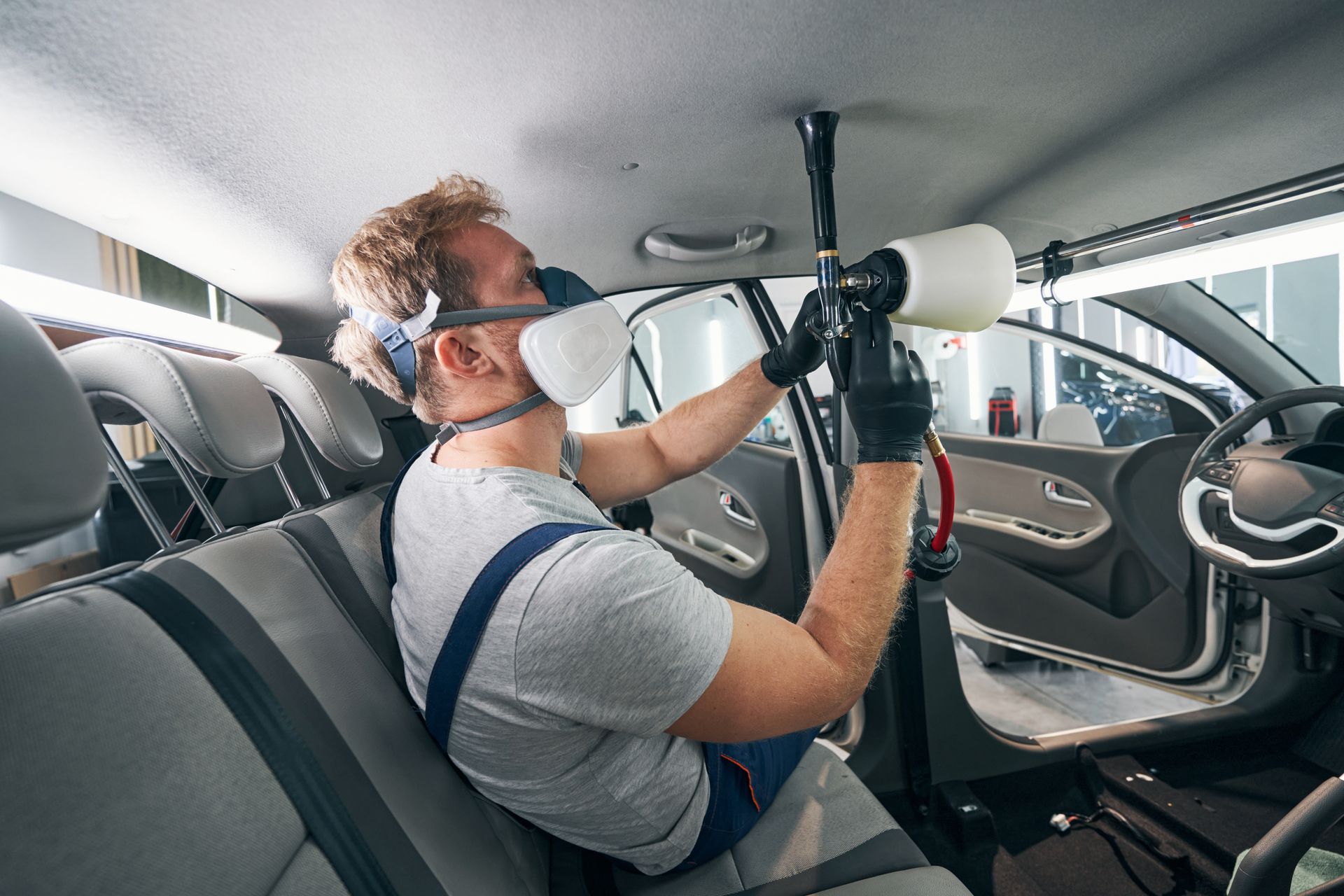 Person cleaning a car's interior with a specialized tool, wearing a respirator, in a car detailing shop.