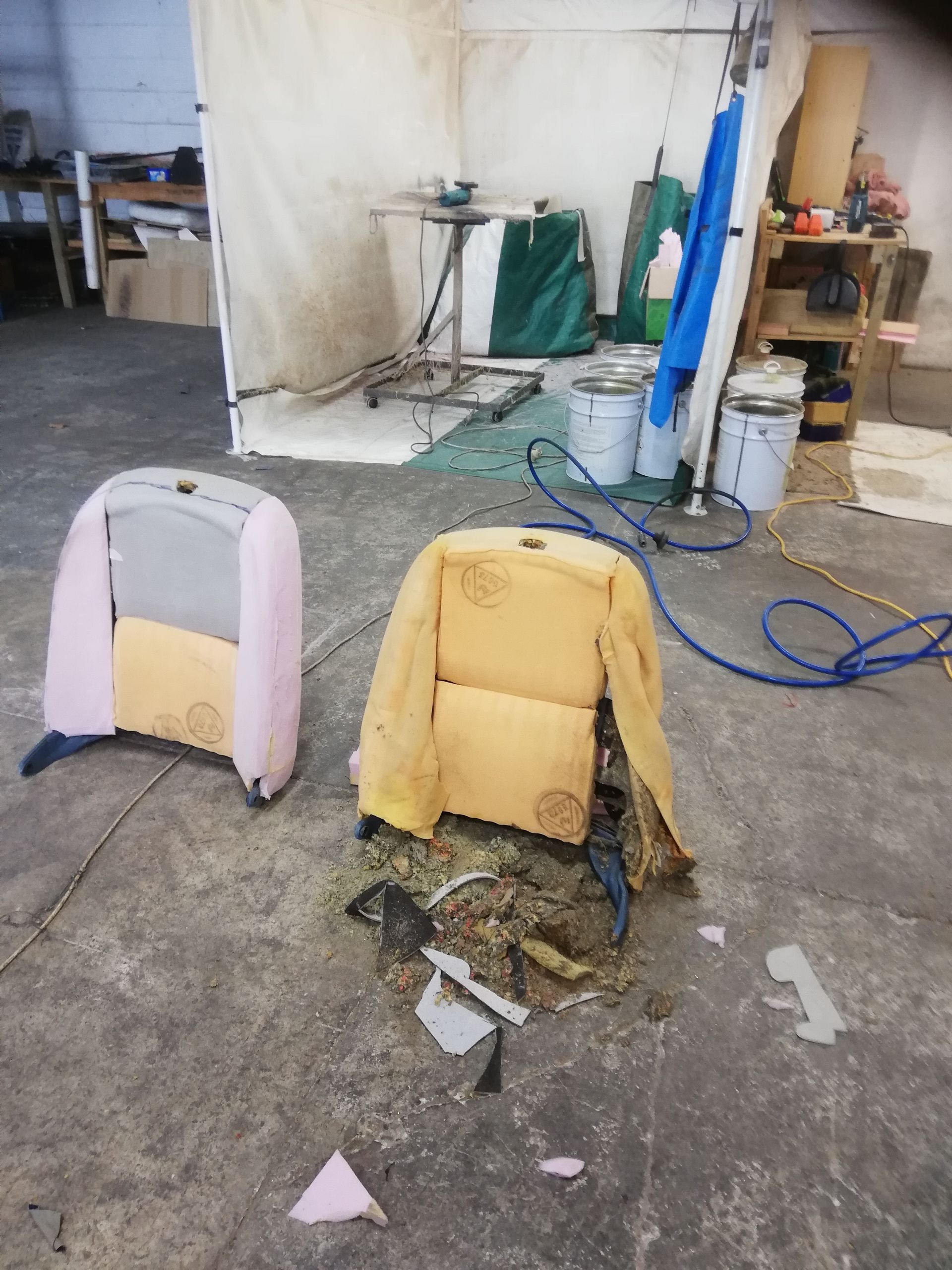Two car seats on a concrete floor. One is pink and intact, the other yellow and torn with debris. Industrial setting.