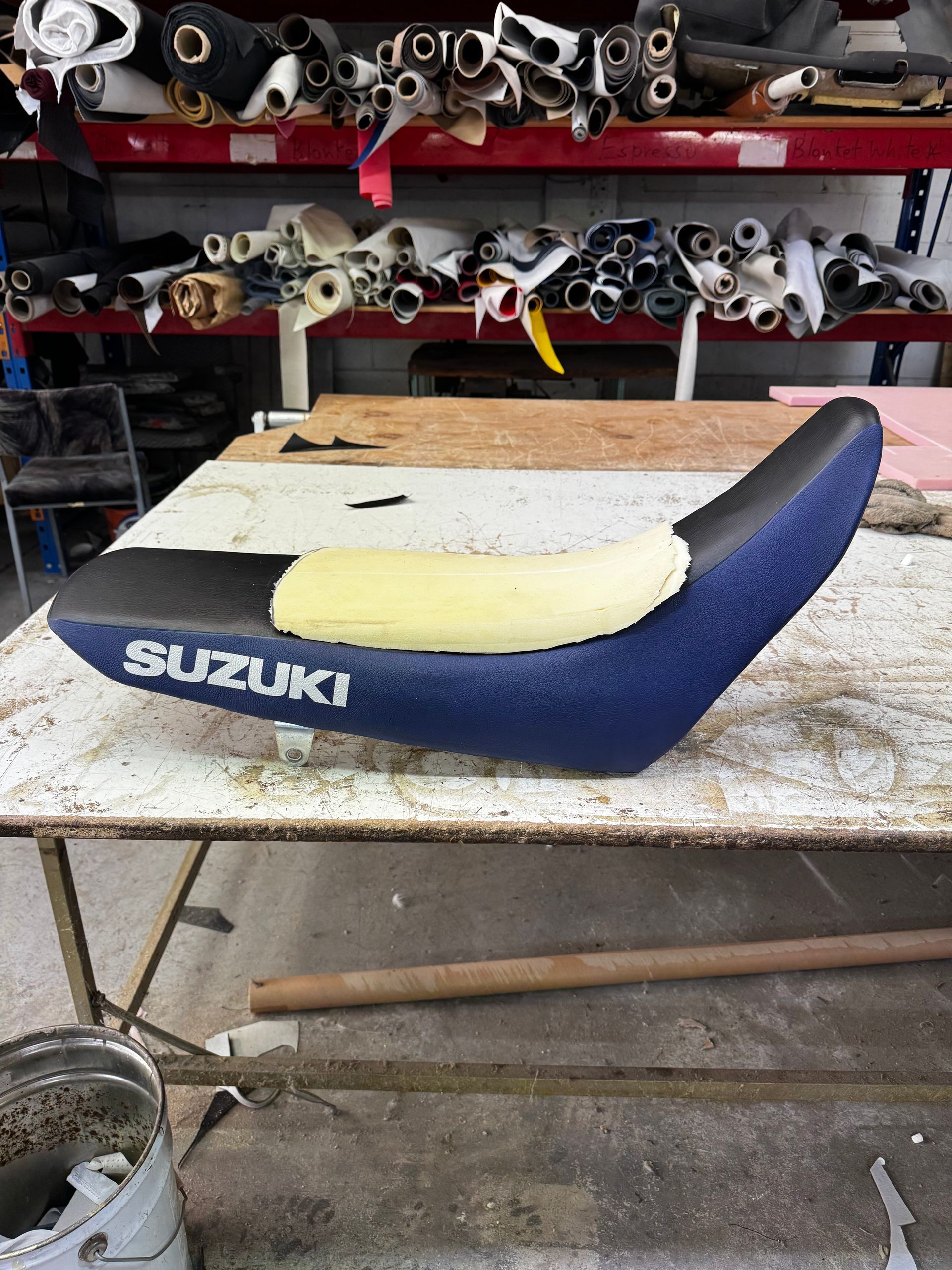 Suzuki motorcycle seat being reupholstered with blue and black material, yellow foam on a workbench.