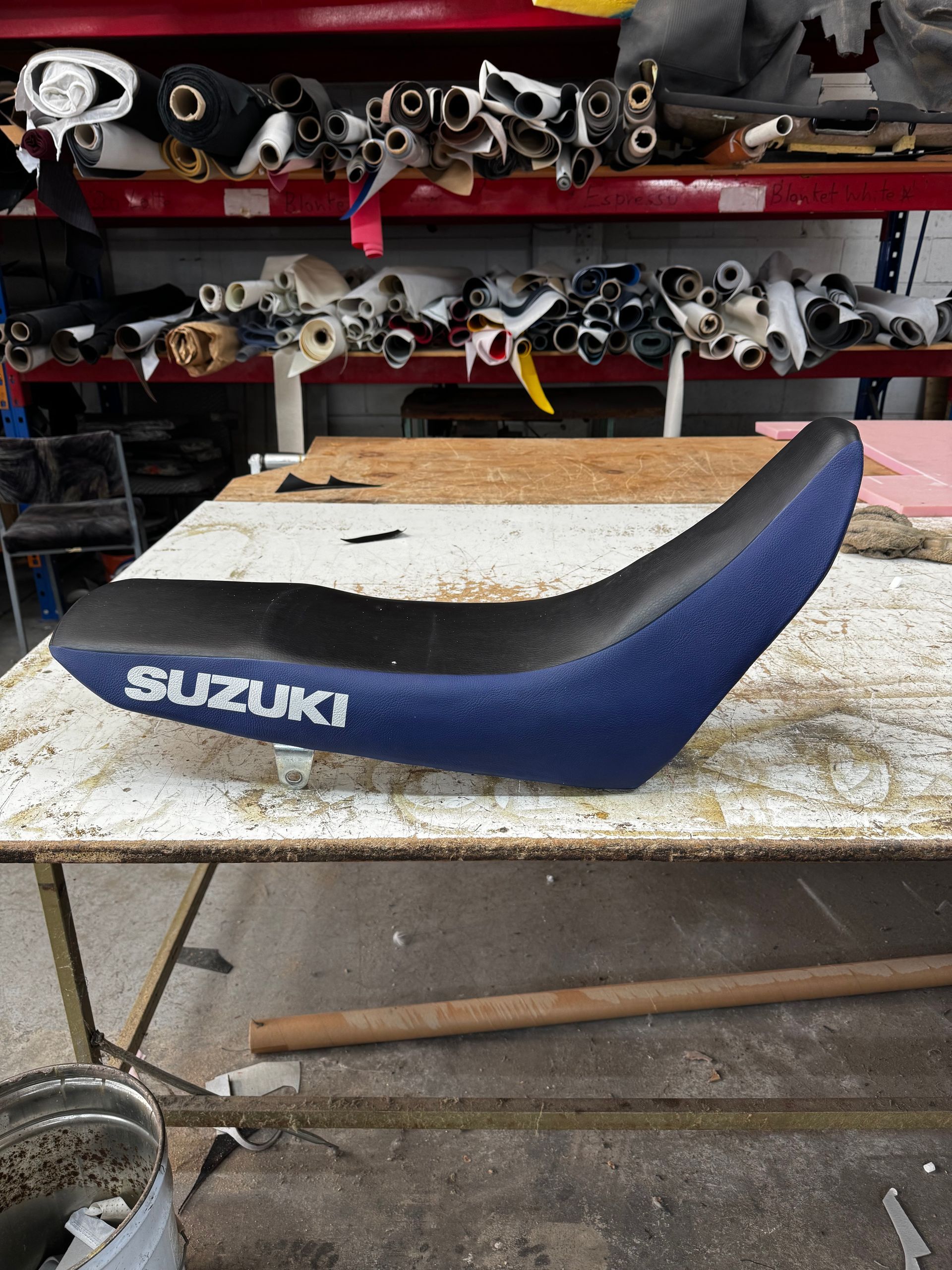 Blue and black Suzuki motorcycle seat on a workbench, fabric rolls in background.