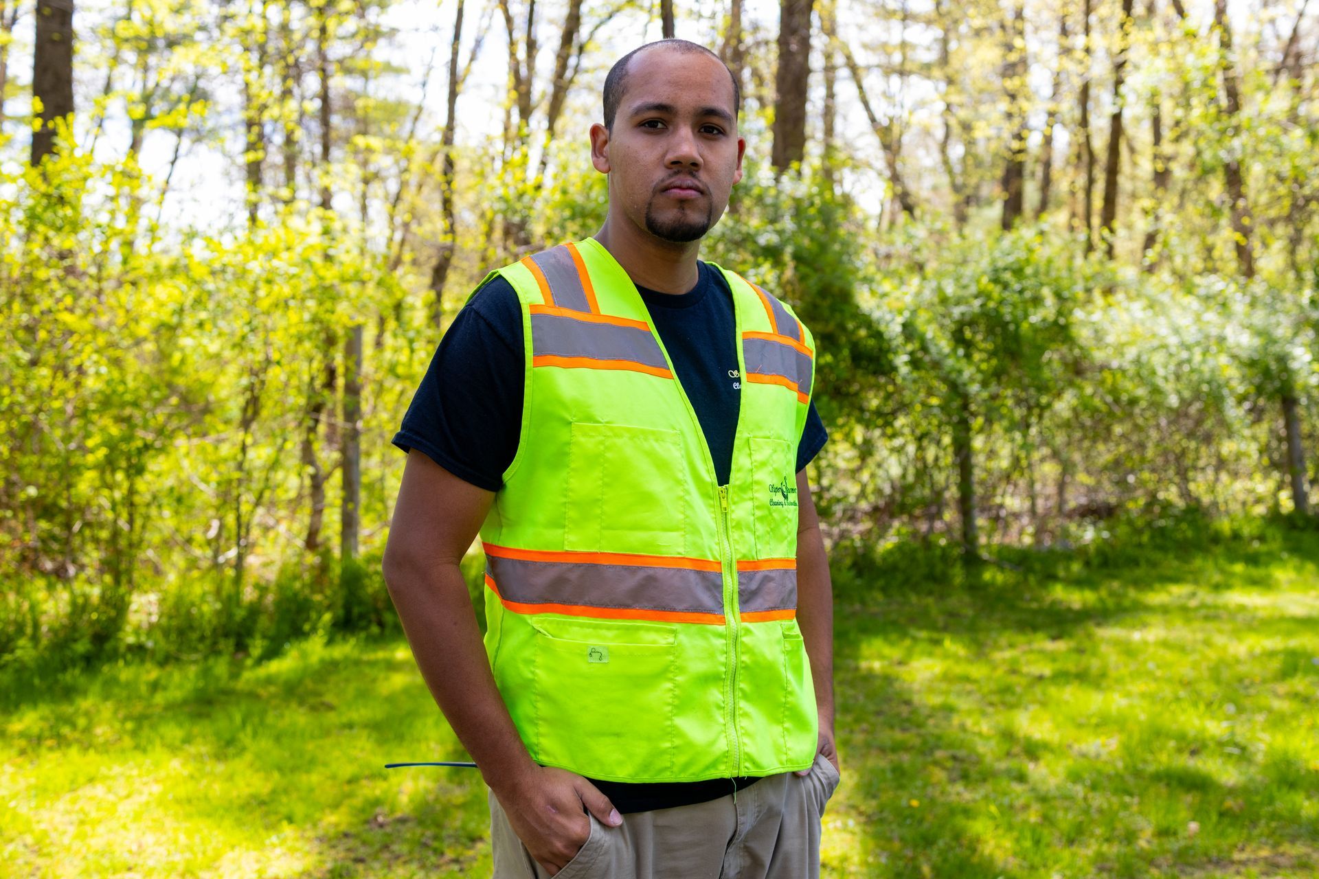 Man wearing a neon green safety vest stands outside with trees in the background.