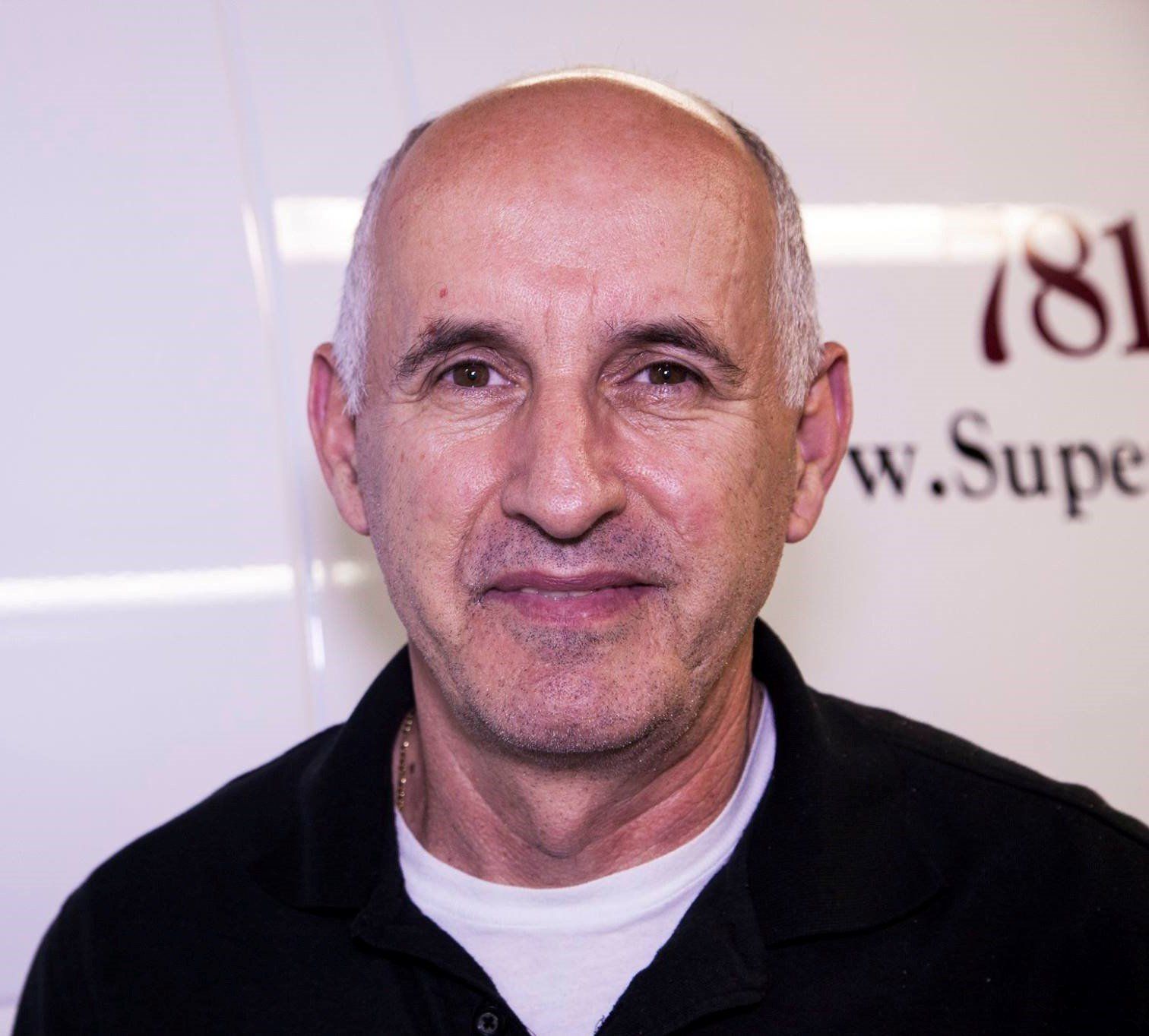 Bald man smiling in front of a white background; wearing a black collared shirt and white undershirt.