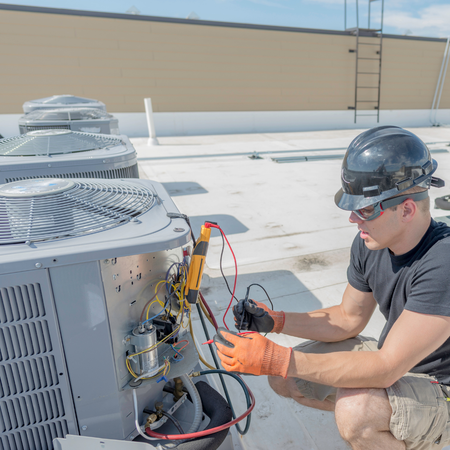 A technician in a hard hat and gloves uses a multimeter to test an HVAC unit on a commercial roof.
