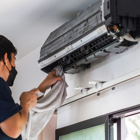A professional wearing a face mask wipes the interior of a wall-mounted air conditioning unit with a cloth.