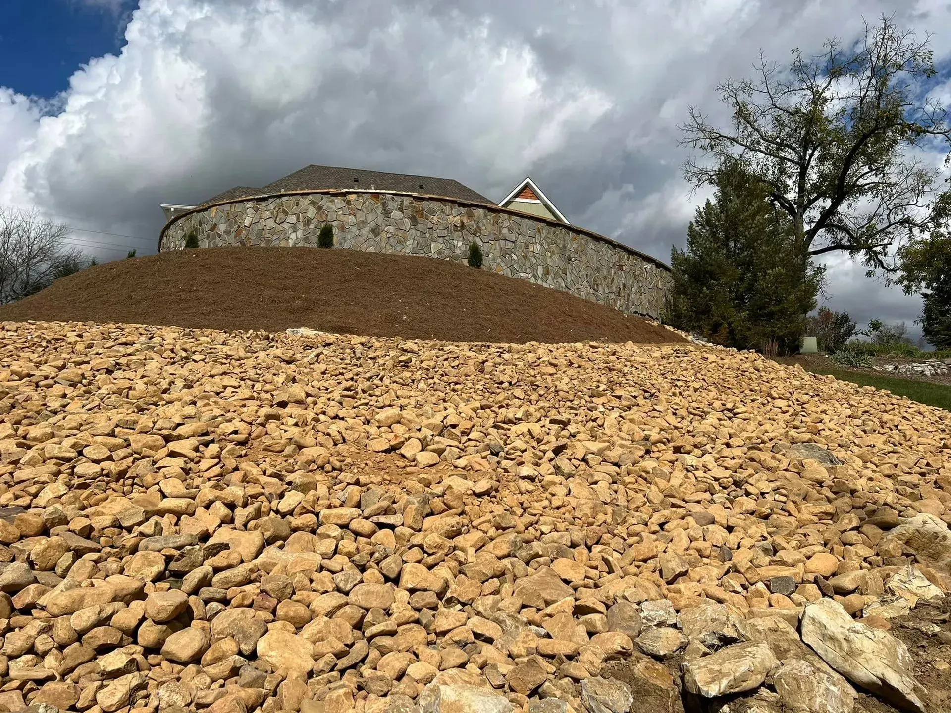 A pile of gravel is sitting on top of a hill with a house in the background.