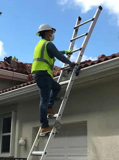 Person in safety gear climbs a ladder to a rooftop. The person wears a hard hat and mask.
