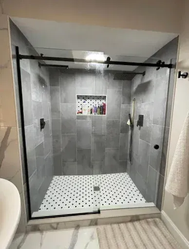 Modern gray tiled shower with glass door and black hardware. Includes a built-in shelf.