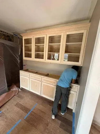 Person installing kitchen cabinets. Upper cabinets with glass doors, lower cabinets with drawers, unfinished wood, and wood flooring.