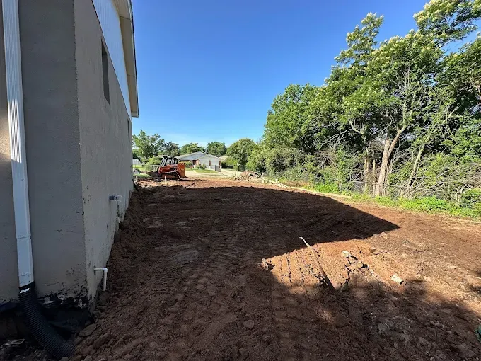 Side view of a house next to a dirt area, with trees in the background and a small construction vehicle.