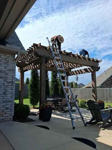 Two people on a ladder working on a wooden pergola, under a blue sky, on a patio.
