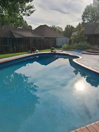 Pool of blue water with brick edging reflecting the sky, surrounded by concrete patio and fences.