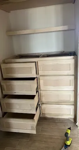 A set of unfinished wooden drawers with open drawers and a shelf inside a closet. A power drill is on the floor.