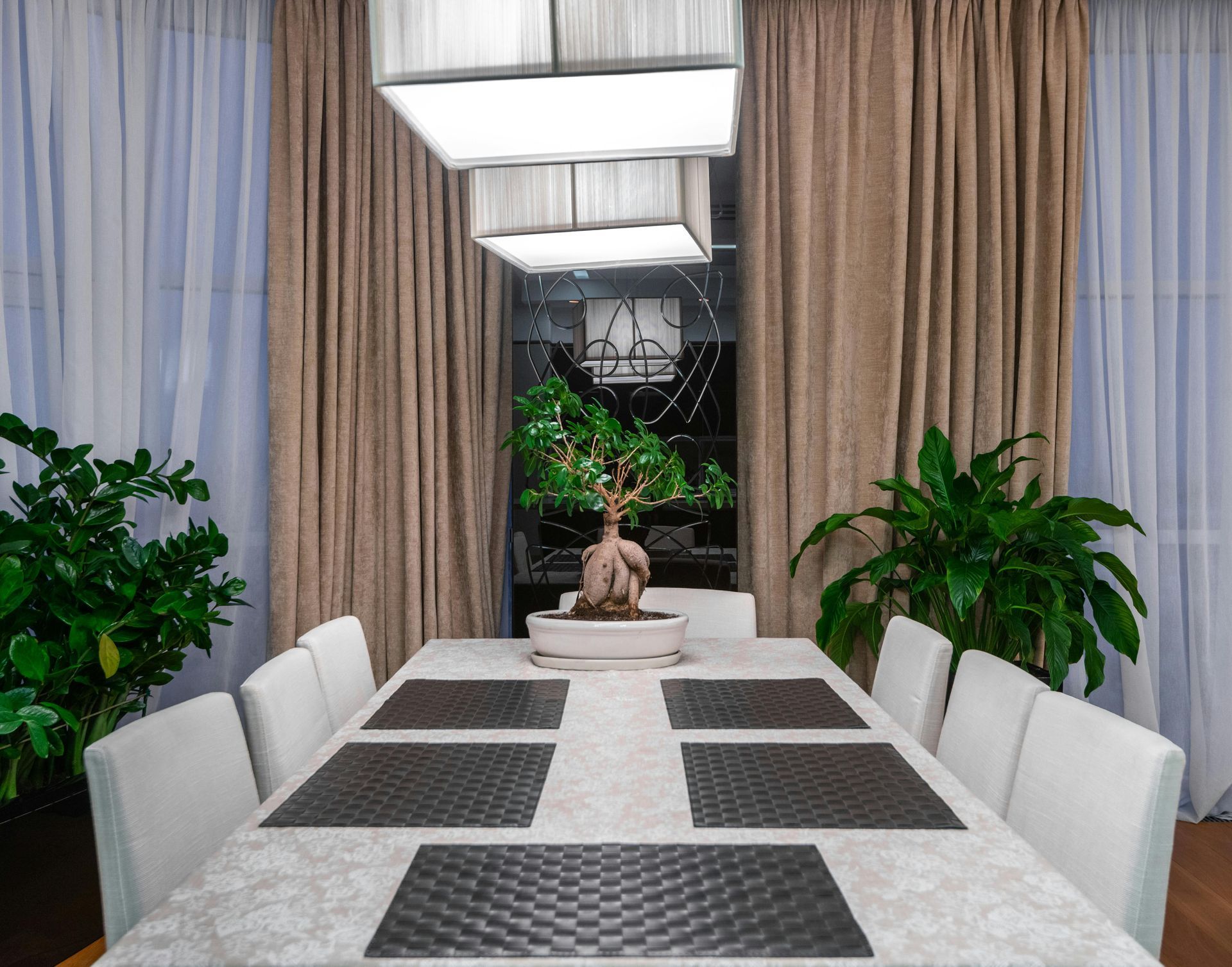 Dining room with a long table, white chairs, plants, and square hanging lights.