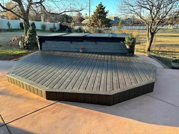 Wooden deck with a hot tub, set on a concrete patio in a backyard.