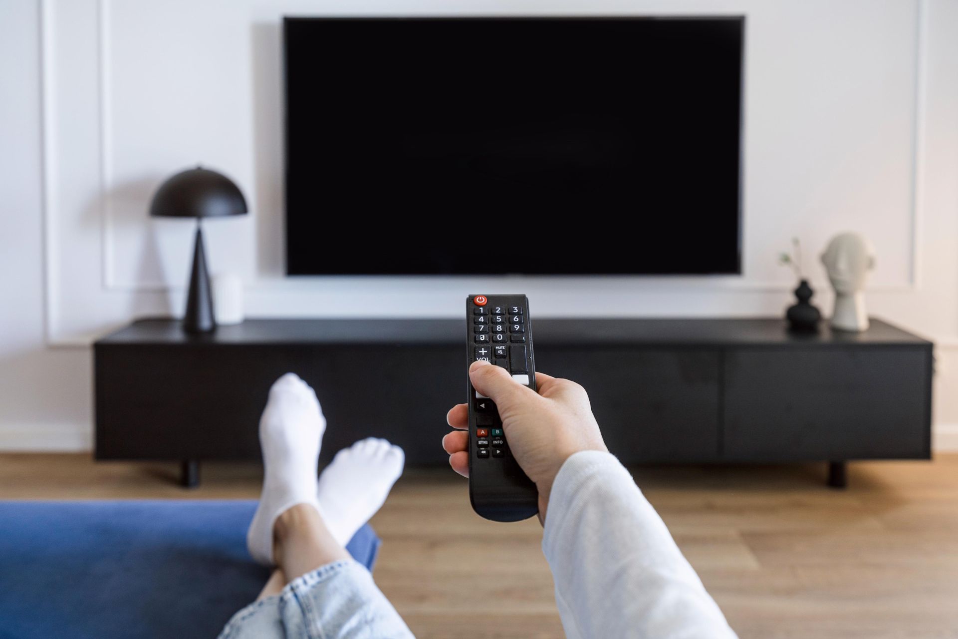 A person is sitting on a couch holding a remote control in front of a television.