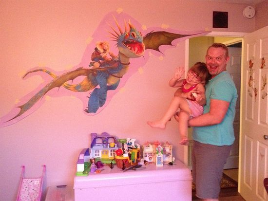 A man is holding a little girl in front of a painting of a dragon on the wall.