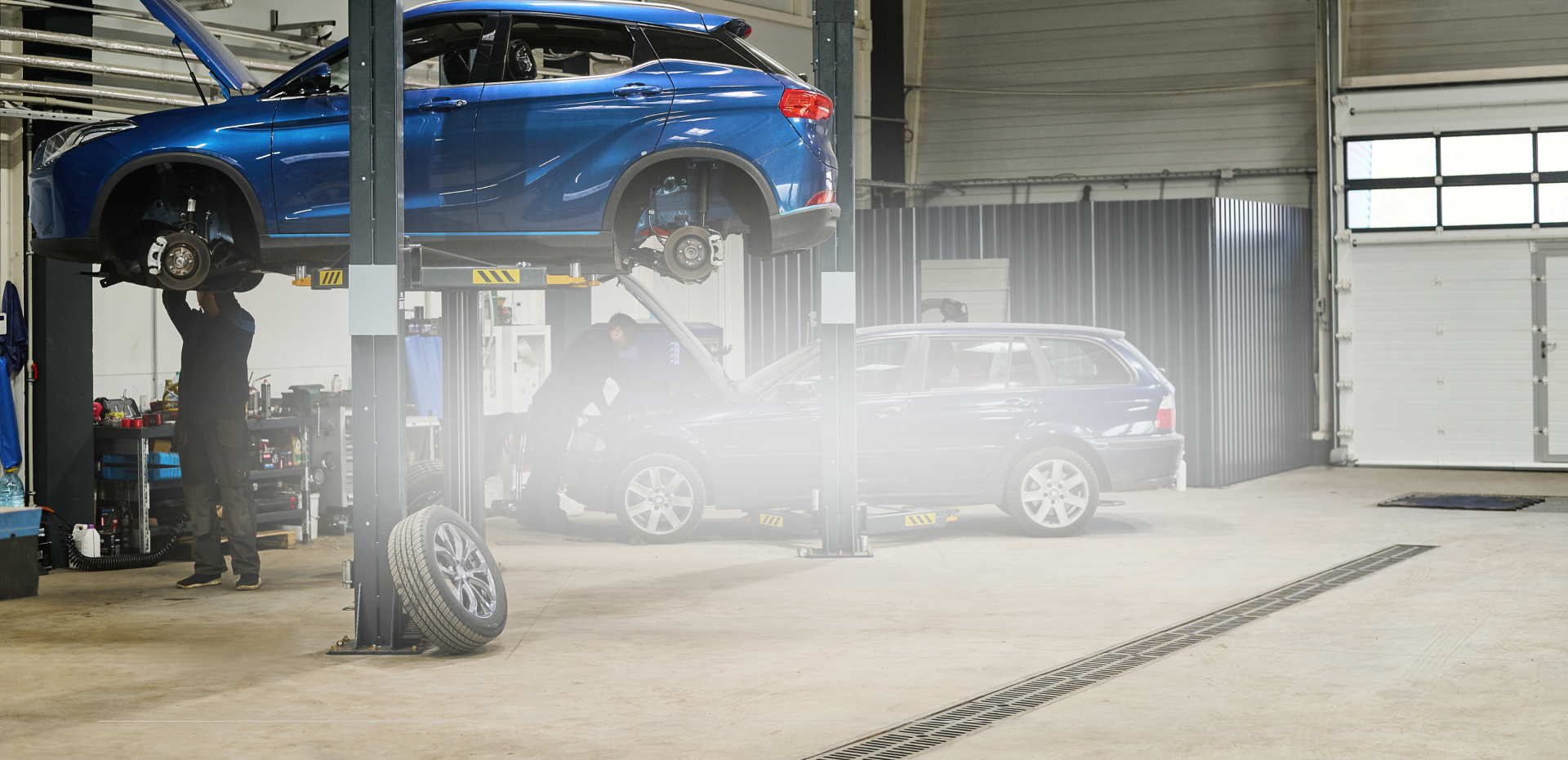 Blue car on a lift in an auto shop, with a silver car below and mist in the garage | Universal Auto Repair
