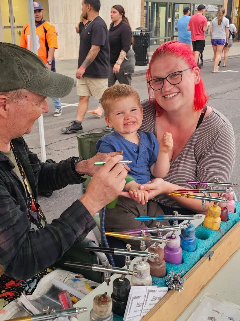 A man is painting a child 's face while a woman holds a baby.