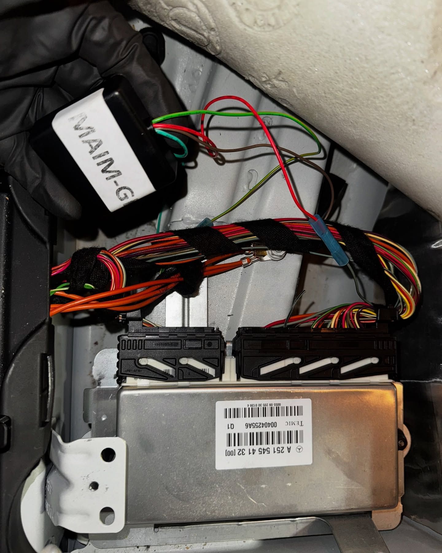 Black-gloved hand holds a black MAIM-G box near car wiring and two electronic control units in a vehicle.
