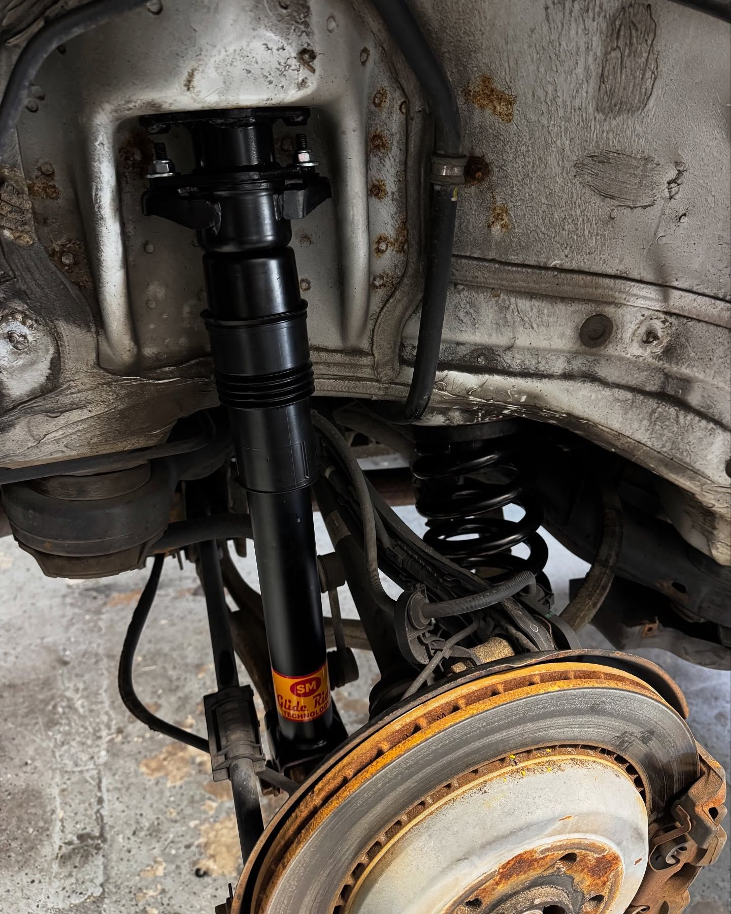 Car's rear suspension being worked on: black shock absorber installed, rusty brake disc, black coil spring.